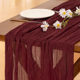 Socomi burgundy red cheesecloth table runner draped on a wedding table for romantic ambiance.